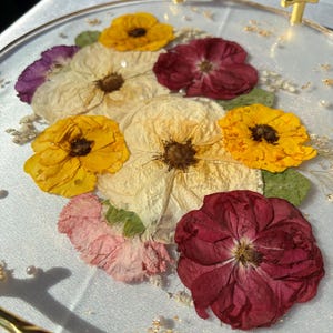 Puede incluir: Una bandeja de servicio redonda y transparente con asas doradas, adornada con flores prensadas en tonos amarillo, crema, rosa y burdeos. Las flores est&aacute;n incrustadas en resina transparente, creando una pieza decorativa y funcional.