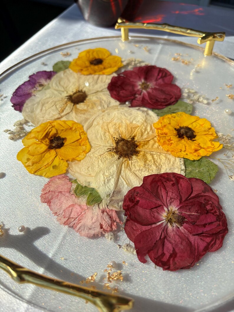 Puede incluir: Una bandeja de servicio redonda y transparente con asas doradas, adornada con flores prensadas en tonos amarillo, crema, rosa y burdeos. Las flores est&aacute;n incrustadas en resina transparente, creando una pieza decorativa y funcional.