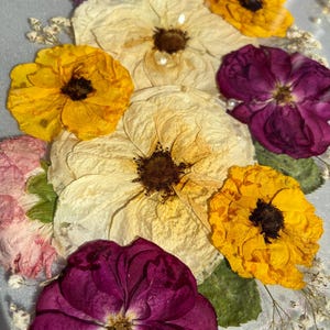 Puede incluir: Primer plano de una bandeja decorativa con flores prensadas. El arreglo incluye flores amarillas, crema y morado oscuro, con delicados acentos blancos. Las flores est&aacute;n sobre un fondo transparente, posiblemente de resina.
