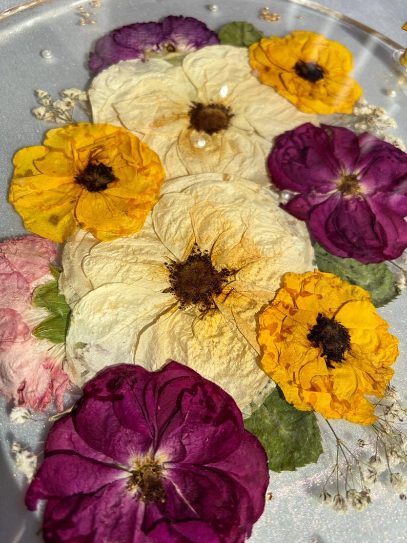 Puede incluir: Primer plano de una bandeja decorativa con flores prensadas. El arreglo incluye flores amarillas, crema y morado oscuro, con delicados acentos blancos. Las flores est&aacute;n sobre un fondo transparente, posiblemente de resina.