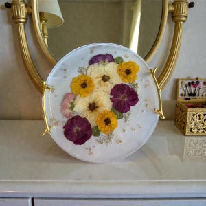 Puede incluir: Un plato decorativo redondo y blanco con un arreglo floral de flores secas amarillas, moradas y crema. El plato tiene asas doradas y detalles de pan de oro.