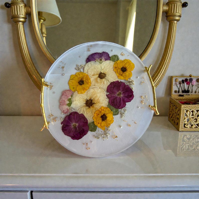Puede incluir: Un plato decorativo redondo y blanco con un arreglo floral de flores secas amarillas, moradas y crema. El plato tiene asas doradas y detalles de pan de oro.