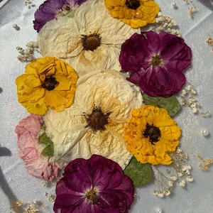 Puede incluir: Una bandeja redonda transparente con un asa dorada, adornada con flores secas en tonos amarillos, morados y crema. El arreglo incluye peque&ntilde;as flores blancas, hojas verdes y detalles de pan de oro, creando un objeto decorativo.