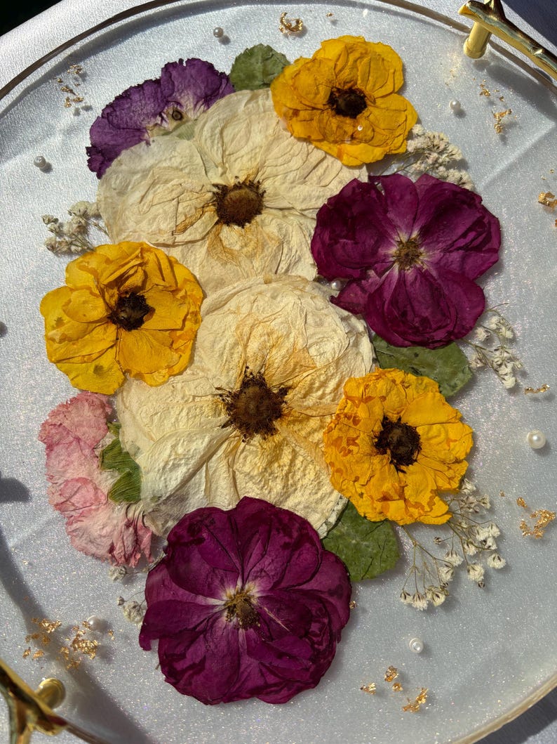Puede incluir: Una bandeja redonda transparente con un asa dorada, adornada con flores secas en tonos amarillos, morados y crema. El arreglo incluye peque&ntilde;as flores blancas, hojas verdes y detalles de pan de oro, creando un objeto decorativo.