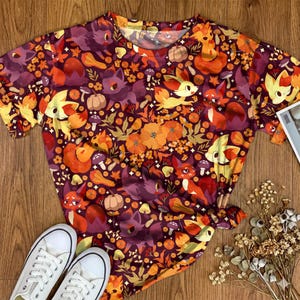 May include: A short-sleeved t-shirt featuring an all-over print of stylised foxes, pumpkins, mushrooms, and orange flowers on a dark purple background. The crew neck shirt displays a repeating pattern of autumn-themed elements and small creatures.
