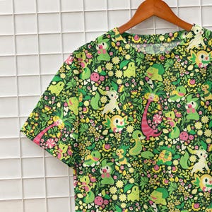 May include: A short-sleeve t-shirt featuring an all-over print of various green and yellow cartoon creatures interspersed with small white and yellow flowers, green leaves, and pink accents on a dark green background, hanging on a wooden hanger.