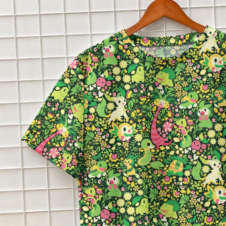 May include: A short-sleeve t-shirt featuring an all-over print of various green and yellow cartoon creatures interspersed with small white and yellow flowers, green leaves, and pink accents on a dark green background, hanging on a wooden hanger.