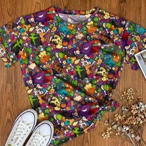 Op de afbeelding: Een t-shirt met korte mouwen en een all-over print van diverse stripfiguren, pompoenen, spoken en groene bladeren op een paarse achtergrond. Het ontwerp bevat kleine schedels, botten en oranje pompoenen die over het kledingstuk verspreid zijn.