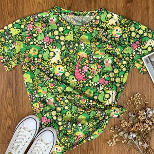 May include: A short-sleeved t-shirt featuring an all-over print of various green, yellow, and pink cartoon creatures interspersed with small flowers, leaves, and vines against a dark green background, with a round neckline and a straight hem.