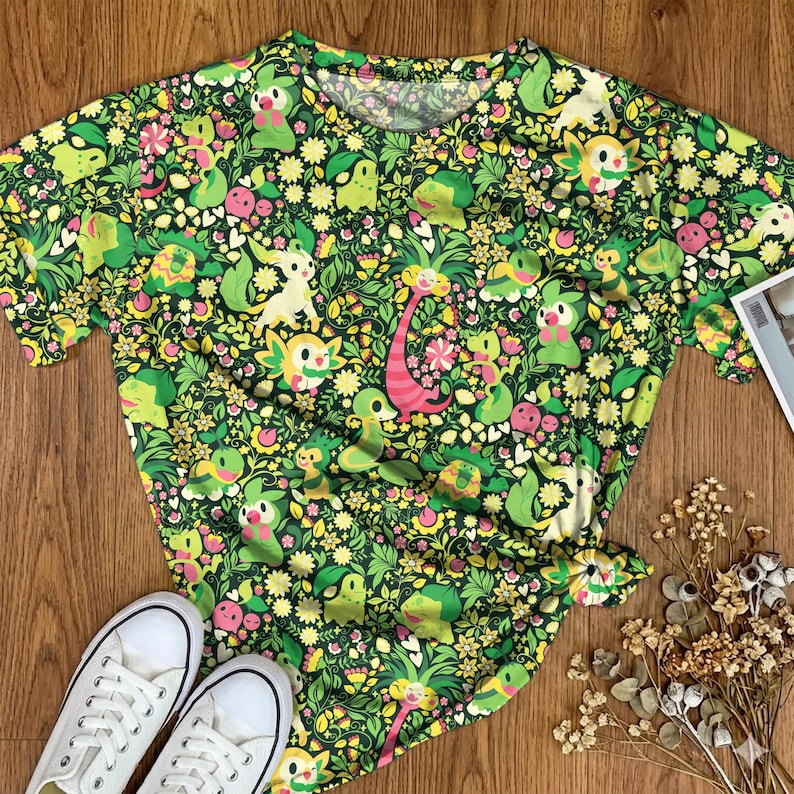 May include: A short-sleeved t-shirt featuring an all-over print of various green, yellow, and pink cartoon creatures interspersed with small flowers, leaves, and vines against a dark green background, with a round neckline and a straight hem.
