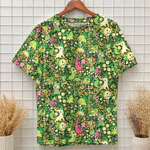May include: A short-sleeved crew neck t-shirt featuring an all-over print of various small green, yellow, and pink cartoon creatures interspersed with yellow flowers, green leaves, and small pink blossoms against a dark green background.