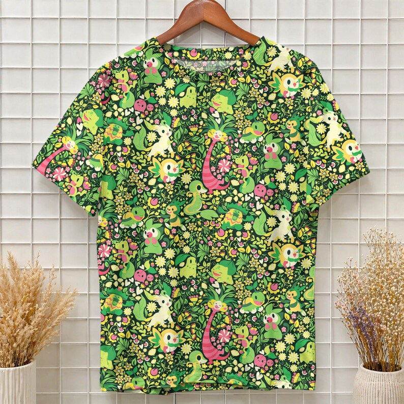 May include: A short-sleeved crew neck t-shirt featuring an all-over print of various small green, yellow, and pink cartoon creatures interspersed with yellow flowers, green leaves, and small pink blossoms against a dark green background.