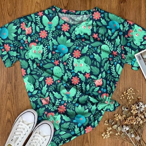 May include: A short-sleeve t-shirt featuring a dark teal background with an all-over pattern of green cartoon creatures, pink flowers, and various leaf shapes. The design includes repeating illustrations of small, light green, and blue characters.