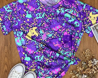 May include: A short-sleeve t-shirt featuring an all-over print with purple, teal, and yellow colors. The graphic pattern includes various stylized ghost-like creatures, skulls, bones, stars, and small hearts scattered across the entire fabric surface.