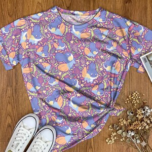 May include: A short-sleeve t-shirt featuring an all-over repeating pattern of blue and orange cartoon creatures, green seaweed, and small yellow stars set against a vibrant purple background with a classic crew neckline and short sleeves.