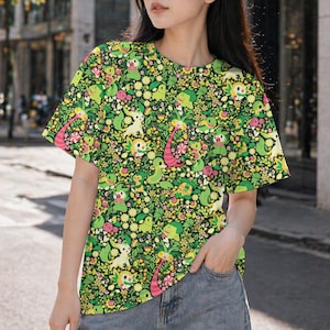 May include: A short-sleeve t-shirt featuring an all-over print of various green, yellow, and pink cartoon creatures interspersed with small flowers and foliage on a dark green background. The shirt has a crew neckline and a straight hemline.