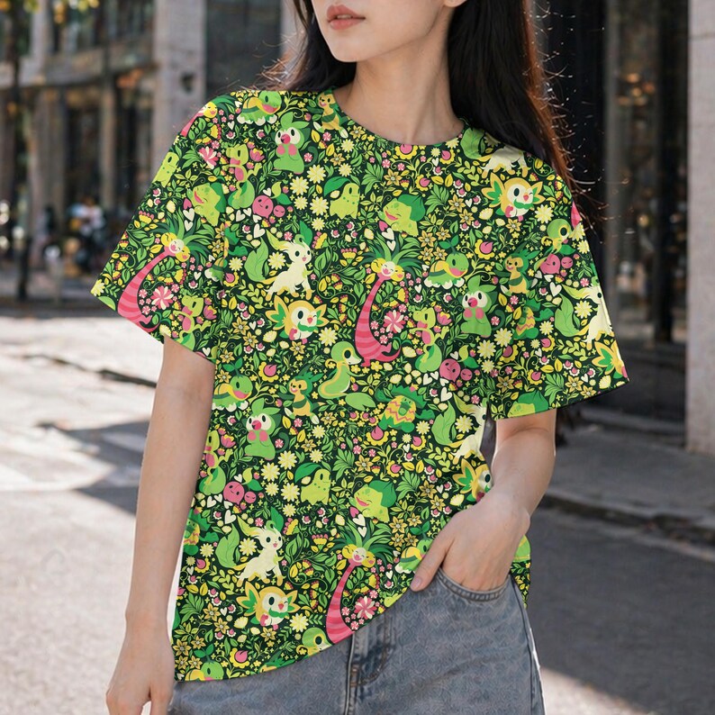 May include: A short-sleeve t-shirt featuring an all-over print of various green, yellow, and pink cartoon creatures interspersed with small flowers and foliage on a dark green background. The shirt has a crew neckline and a straight hemline.