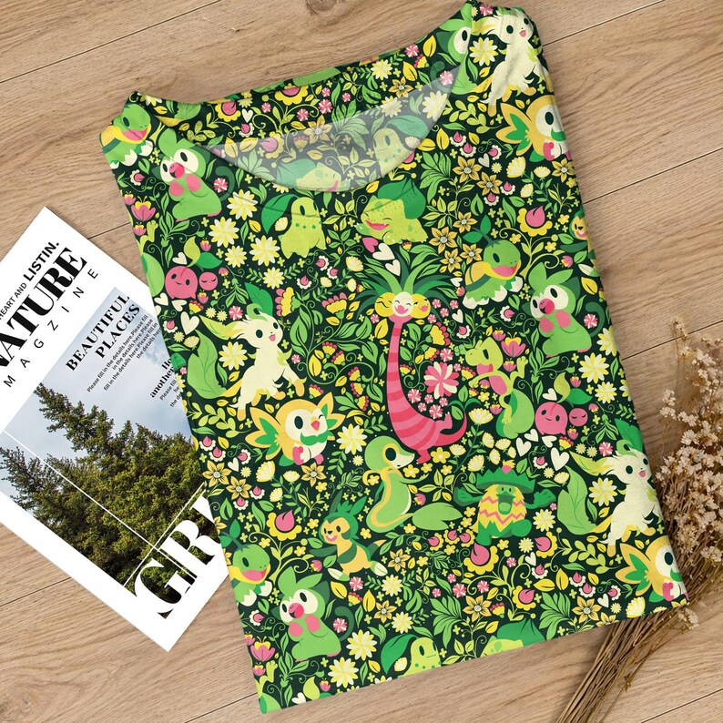 May include: A short-sleeved t-shirt featuring an all-over print of various green, yellow, and pink cartoon creatures interspersed with small yellow and pink flowers and white heart shapes against a dark green background with a round neckline.