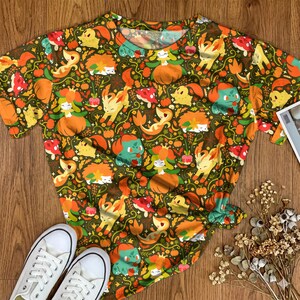Op de afbeelding: Een t-shirt met korte mouwen en een all-over print van diverse cartoonfiguren, oranje pompoenen en groene ranken op een donker olijfgroene achtergrond. Het patroon bevat herhalende illustraties van kleine gele, oranje en groene karakters.