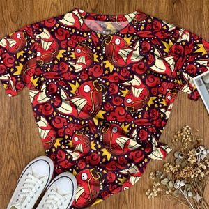 May include: A short-sleeved t-shirt featuring an all-over print of red fish with cream-coloured fins, yellow star shapes, and dark red swirling cloud patterns against a deep purple background. The shirt has a round neckline and short sleeves.