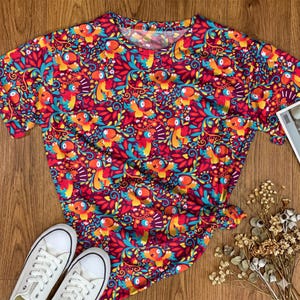 Può includere: Una t-shirt a maniche corte con un vivace motivo ripetuto di pappagalli colorati, foglie stilizzate color ottanio e piccoli fiori bianchi su uno sfondo magenta intenso. La maglietta presenta un classico scollo rotondo.