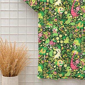 May include: A short-sleeved t-shirt featuring a dense, repeating pattern of various green and yellow cartoon creatures interspersed with small pink and white flowers, green leaves, and pink heart shapes against a dark green background.