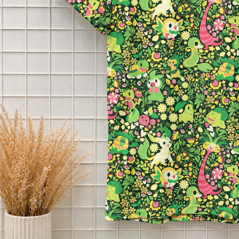 May include: A short-sleeved t-shirt featuring a dense, repeating pattern of various green and yellow cartoon creatures interspersed with small pink and white flowers, green leaves, and pink heart shapes against a dark green background.