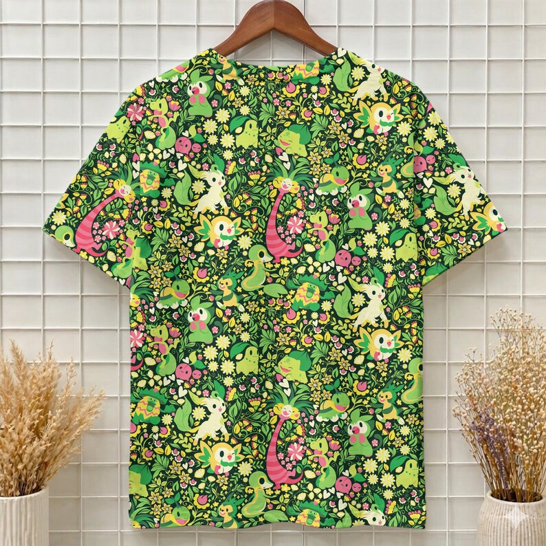 May include: A short-sleeve t-shirt featuring an all-over print of various green, yellow, and pink cartoon creatures interspersed with small flowers, leaves, and hearts on a dark green background, displayed on a wooden hanger.
