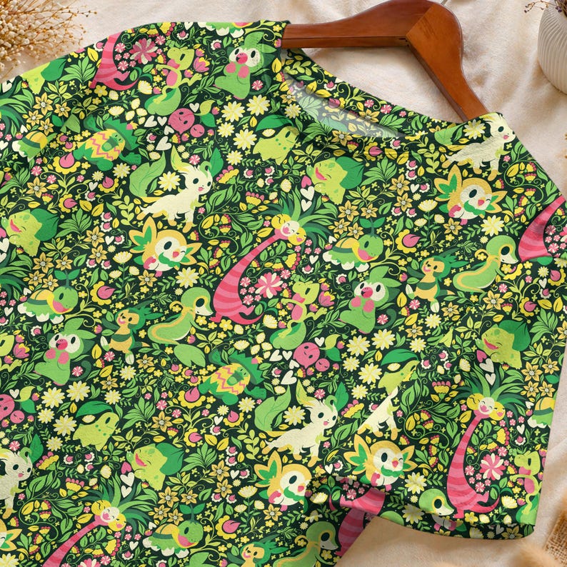May include: A short-sleeved t-shirt featuring an all-over print of various green-toned cartoon creatures interspersed with yellow and pink flowers, green leaves, and small white hearts against a dark green background on a wooden hanger.