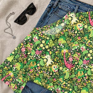 May include: A short-sleeve t-shirt featuring an all-over print of various green and yellow cartoon creatures interspersed with small flowers, leaves, and hearts on a dark green background, paired with blue denim jeans, black sunglasses, and a silver chain.