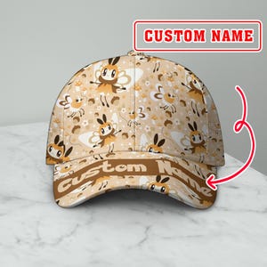 Peut inclure: Casquette de baseball beige avec un motif répété d'abeilles, de fleurs et de champignons de dessin animé. La visière et le texte "CUSTOM NAME" sont marron foncé. Le texte "CUSTOM NAME" est également sur le devant de la casquette.