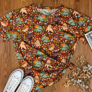 Op de afbeelding: Een t-shirt met korte mouwen en een all-over print met diverse stripfiguren, pompoenen, zonnebloemen en het woord BOO in een herhalend patroon. Het ontwerp heeft een donkerbruine achtergrond met oranje, groene en gele accenten.