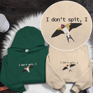 May include: A dark green hoodie and a beige sweatshirt, both featuring an embroidered bird design with the text "I don't spit, I". The bird has black, yellow, red, and white details. The hoodie has a hood, and the sweatshirt has long sleeves.