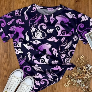 May include: A short-sleeved t-shirt featuring a dark navy blue background with a repeating pattern of stylized purple and white cat-like creatures and swirling cloud shapes. The design covers the entire garment, including the crew neckline.