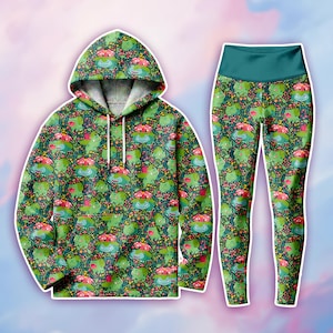 Op de afbeelding: Een bijpassende set bestaande uit een sweatshirt met lange mouwen en capuchon met een kangoeroezak en een legging met hoge taille. Beide kledingstukken hebben een herhalend patroon van groene wezens met roze bloemknoppen op een donkere achtergrond.