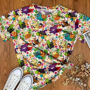 Könnte beinhalten: Ein kurzärmeliges T-Shirt mit einem Allover-Druck aus verschiedenen Zeichentrickfiguren, Kürbislaternen und Geistern in leuchtenden Farben wie Lila, Orange, Grün und Gelb auf einem weißen Hintergrund mit kleinen floralen Akzenten.