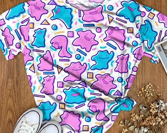 May include: A white short-sleeve t-shirt featuring an all-over pattern of pink and blue blob-shaped characters with simple smiling faces, interspersed with small yellow triangles, blue rectangles, and circles on a light grid background.