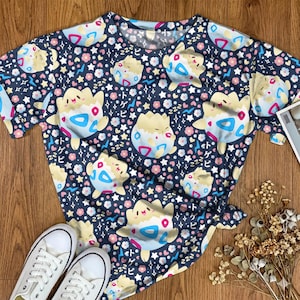 May include: Navy blue short-sleeved t-shirt featuring a repeating pattern of cartoon characters, flowers, stars, and Japanese text. The shirt is paired with white canvas sneakers. The overall theme is playful and whimsical.