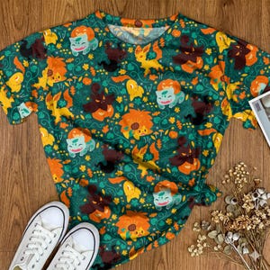Op de afbeelding: Een t-shirt met korte mouwen en een all-over print op een donkergroene achtergrond. Het patroon bevat illustraties van lichtgroene wezens, oranje pompoenen, gele vosachtige figuren, bruine spookachtige vormen, spinnenwebben en paddenstoelen.