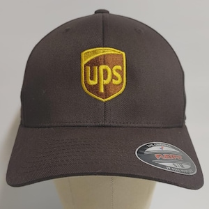 May include: A brown baseball cap with a gold and brown UPS logo embroidered on the front. The cap has a black Flexfit label on the back.