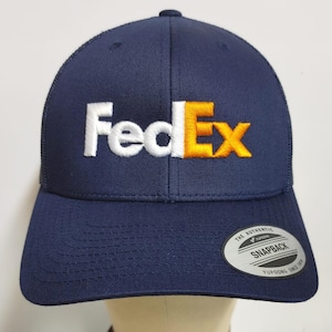 May include: A navy blue trucker hat with a white and orange embroidered FedEx logo. The hat has a black snap closure and a white "The Authentic Yupoong Snapback" label on the back.