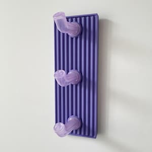 Könnte beinhalten: Ein lilafarbener Wandgarderobenständer mit drei gebogenen, durchscheinenden lavendelfarbenen Haken. Der Ständer hat eine rechteckige Form mit vertikalen Rippendetails. Die Haken sind gleichmäßig verteilt und ragen aus dem Ständer heraus.