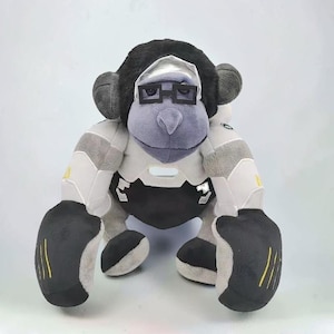 Peut inclure: Une peluche de gorille avec un visage gris, des poils noirs sur la tête et une combinaison de style robotique blanche, grise et noire. Le gorille porte des lunettes rectangulaires noires et un bandeau argenté sur le front.