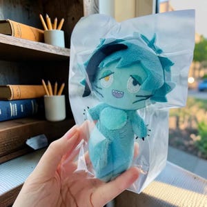 Puede incluir: Un peluche de color verde azulado con peinado puntiagudo, ojos amarillos y una boca pequeña con dientes visibles, dentro de una bolsa de plástico transparente. El personaje tiene marcas oscuras en el cabello y la cara, con extremidades pequeñas.