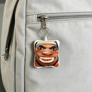 May include: A square-shaped fabric keyring featuring a printed illustration of a person with a wide, toothy grin and a tan headband. The keyring is attached to the silver metal zip pull of a light grey fabric backpack with a visible zip track.