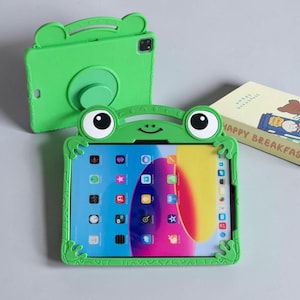 Könnte beinhalten: Eine leuchtend grüne Tablet-Hülle in Froschform mit großen Augen und einem lächelnden Mund. Die Hülle verfügt über einen integrierten Ständer auf der Rückseite und umschließt ein Tablet, das einen bunten Startbildschirm mit App-Symbolen zeigt.