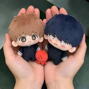 Puede incluir: Dos pequeñas muñecas de peluche con ojos grandes y pelo suave, una con pelo castaño claro y la otra con pelo azul oscuro, ambas con trajes azul marino. Un diminuto peluche de pulpo rojo brillante se encuentra entre las dos muñecas.