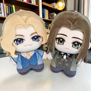 Pode incluir: Dois bonecos de pelúcia com grandes olhos bordados e cabelo detalhado. O boneco da esquerda tem cabelo loiro, jaqueta azul claro e colete estampado. O boneco da direita tem cabelo castanho, jaqueta verde suave e colete escuro estampado.