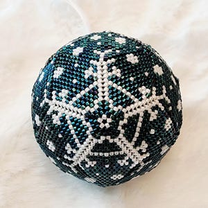 May include: A beaded Christmas ornament in the shape of a sphere. The ornament is primarily dark teal with a white snowflake design. The ornament has a silver string attached for hanging.