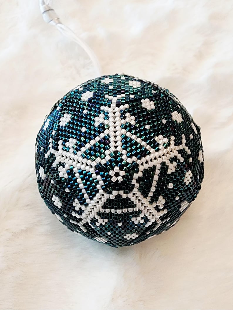 May include: A beaded Christmas ornament in the shape of a sphere. The ornament is primarily dark teal with a white snowflake design. The ornament has a silver string attached for hanging.